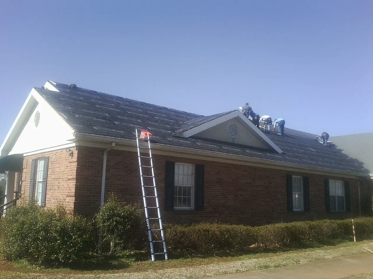 Crew tearing shingles off a brick ranch roof with a ladder set — Metal Roof Repair work by our crew in Penney Farms
