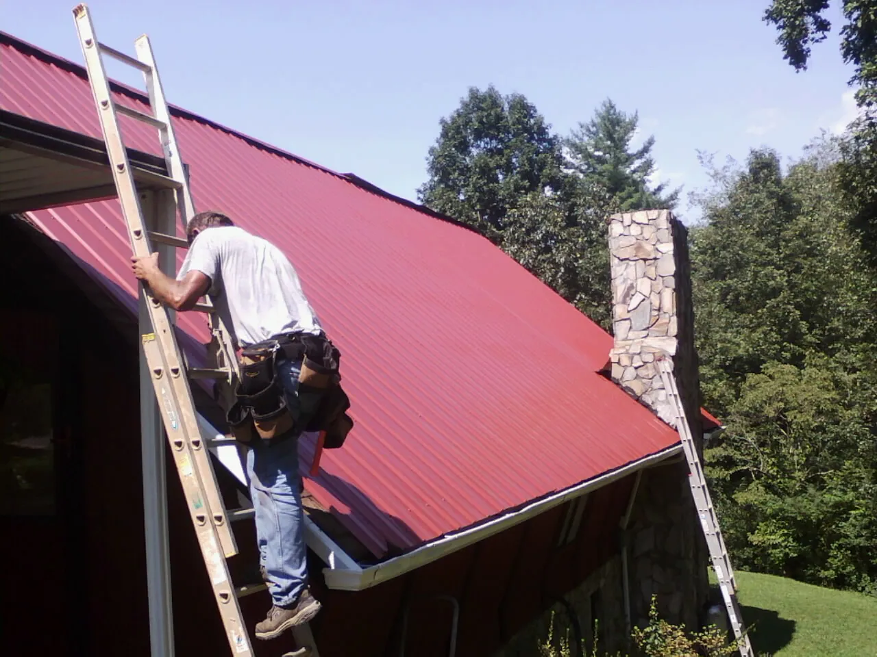 Roofer on a ladder beside a red metal roof and stone chimney — Soffit & Fascia Repair by Roofing Brothers in Penney Farms