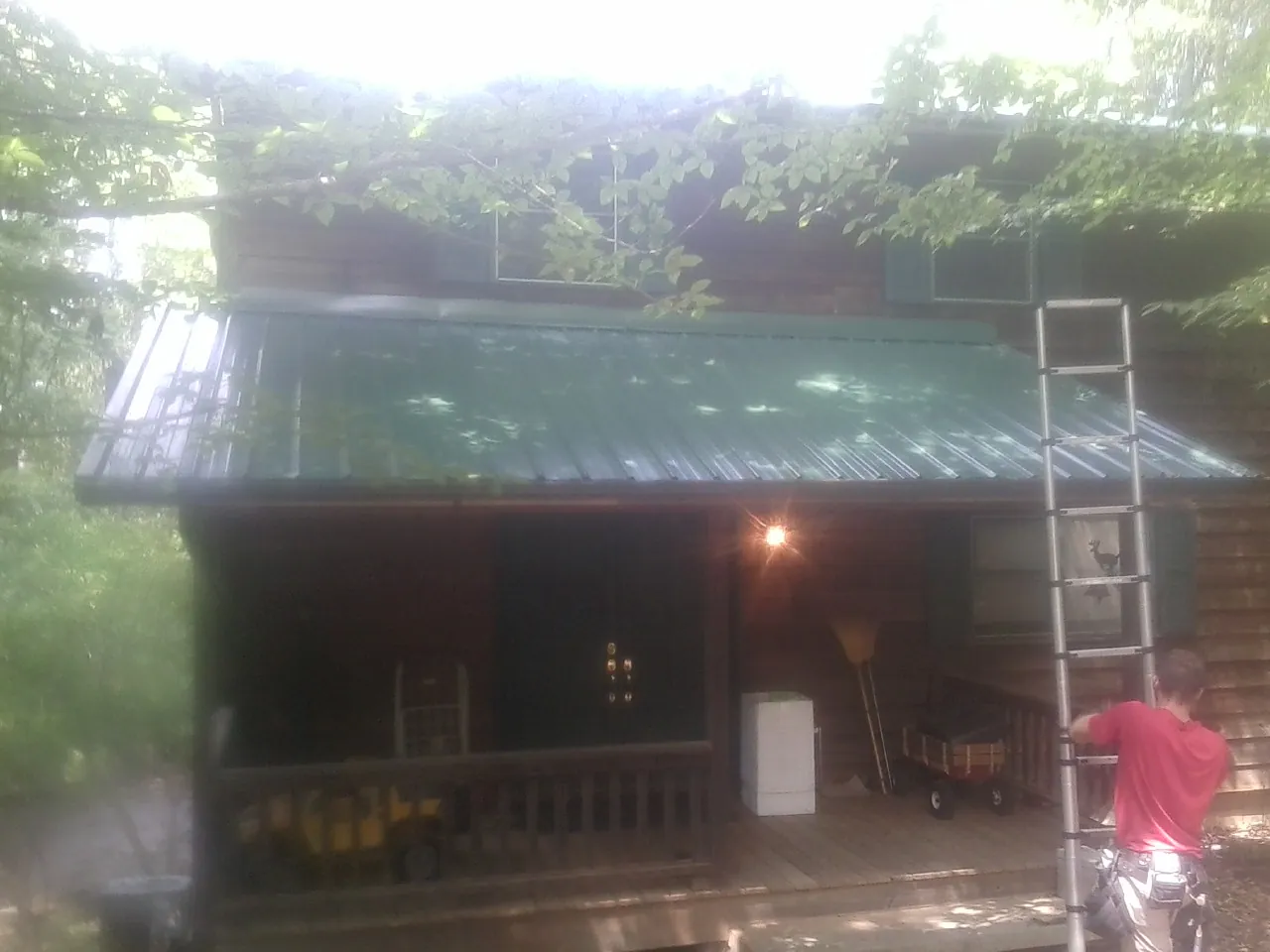 Worker on a ladder at the green porch roof of a wooded cabin in Penney Farms
